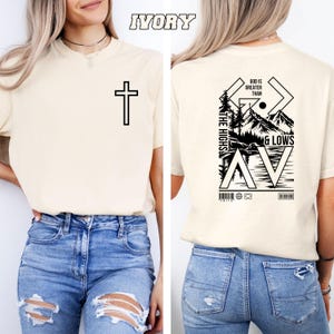 May include: Ivory-colored t-shirt with a black cross on the front. The back of the shirt features a graphic with the text "GOD IS GREATER THAN THE HIGHS & LOWS" and a mountain scene. The shirt is made of a soft material.