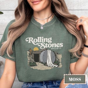 May include: A green t-shirt with a vintage-style graphic of a stone tomb with a white sheet draped over it. The text "Rolling Stones Since AD 33" is printed on the shirt.
