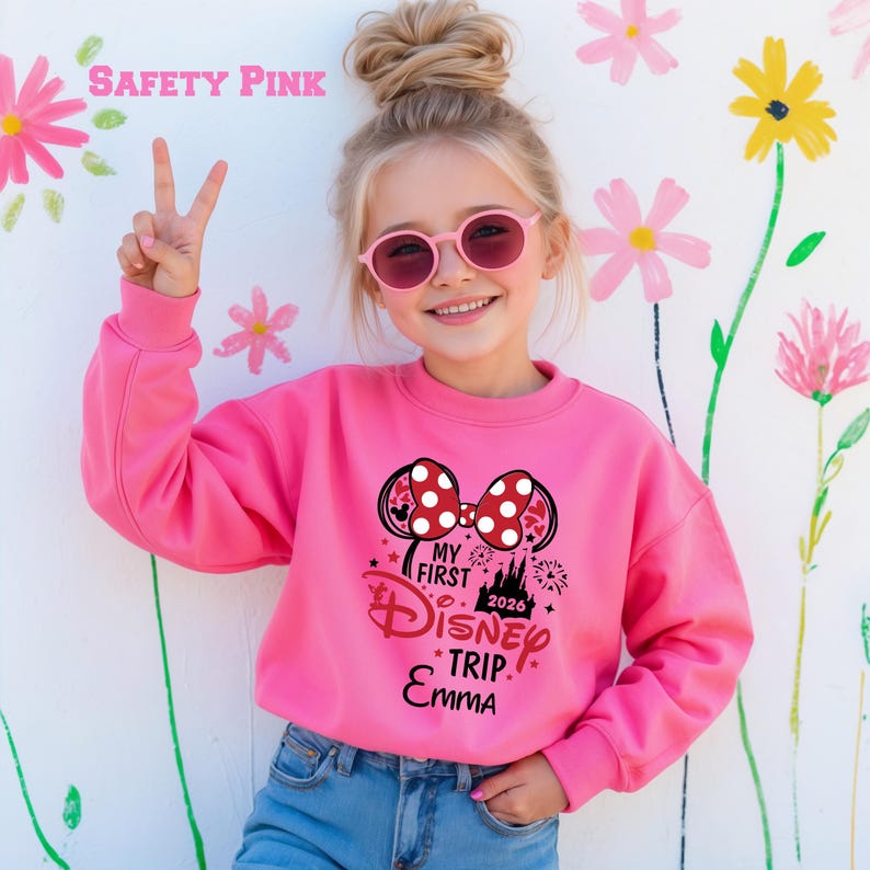 May include: A Safety Pink sweatshirt with a Minnie Mouse bow design and the text "My First Disney Trip Emma 2026". The sweatshirt is worn by a young girl wearing pink sunglasses and blue jeans.