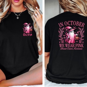 May include: Black t-shirt with a pink ghost wearing a witch hat and holding a pumpkin. The text "In October We Wear Pink" and "Breast Cancer Awareness" is printed on the back. The front of the shirt has a pink ghost with a witch hat and the name "Olivia" printed below.