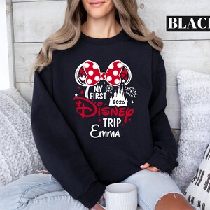 May include: Black sweatshirt with a Minnie Mouse bow graphic. The graphic includes the text "MY FIRST Disney TRIP 2026 Emma" in red and white. The word "BLACK" is printed in a white rectangle at the top.