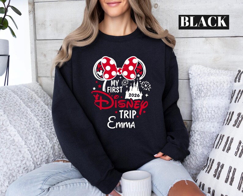 May include: Black sweatshirt with a Minnie Mouse bow graphic. The graphic includes the text "MY FIRST Disney TRIP 2026 Emma" in red and white. The word "BLACK" is printed in a white rectangle at the top.