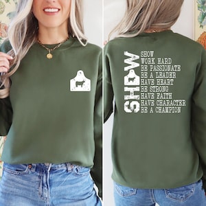 May include: An olive green sweatshirt with a white graphic of a cow tag on the front. The back of the sweatshirt features the word "SHOW" vertically, along with inspirational text: "WORK HARD, BE PASSIONATE, BE A LEADER..."