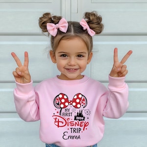 May include: A light pink sweatshirt with a Minnie Mouse design, featuring a red and white polka dot bow and the text "MY FIRST DISNEY TRIP. EMMA 2026". The girl is wearing pink bows in her hair and making a peace sign.