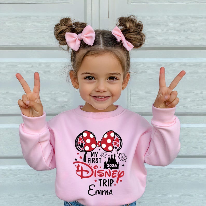 May include: A light pink sweatshirt with a Minnie Mouse design, featuring a red and white polka dot bow and the text "MY FIRST DISNEY TRIP. EMMA 2026". The girl is wearing pink bows in her hair and making a peace sign.