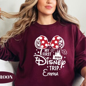 May include: Maroon sweatshirt with a white graphic featuring a Minnie Mouse bow with red polka dots, the text "My First Disney Trip 2026 Emma", and Disney-themed elements. The sweatshirt is a solid maroon color.