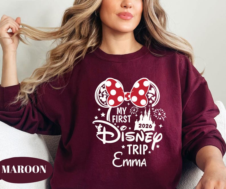 May include: Maroon sweatshirt with a white graphic featuring a Minnie Mouse bow with red polka dots, the text "My First Disney Trip 2026 Emma", and Disney-themed elements. The sweatshirt is a solid maroon color.