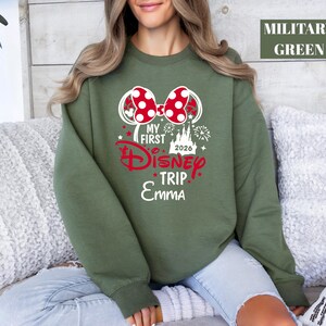 May include: Military green sweatshirt with a graphic that says "My First Disney Trip 2026 Emma". The graphic includes a red bow, a castle, and fireworks. The sweatshirt is long-sleeved.