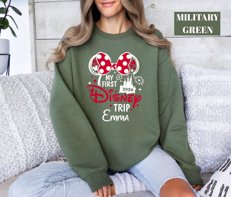 May include: Military green sweatshirt with a graphic that says "My First Disney Trip 2026 Emma". The graphic includes a red bow, a castle, and fireworks. The sweatshirt is long-sleeved.