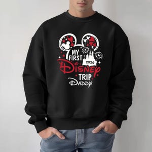 May include: Black sweatshirt with a Disney-themed graphic. The design includes Mickey Mouse ears, fireworks, stars, and the text "MY FIRST Disney TRIP DADDY 2026" in red and white.
