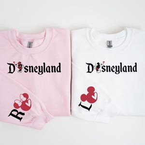 Puede incluir: Dos sudaderas, una rosa y otra blanca. Cada una lleva la palabra "Disneyland" en escritura negra, con un dise&ntilde;o de Mickey y Minnie Mouse. La sudadera rosa tiene un dise&ntilde;o de Minnie Mouse roja y la letra "R" en la manga. La blanca tiene la letra "L".
