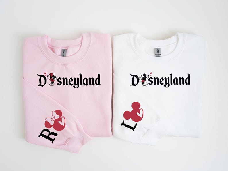 Puede incluir: Dos sudaderas, una rosa y otra blanca. Cada una lleva la palabra "Disneyland" en escritura negra, con un dise&ntilde;o de Mickey y Minnie Mouse. La sudadera rosa tiene un dise&ntilde;o de Minnie Mouse roja y la letra "R" en la manga. La blanca tiene la letra "L".
