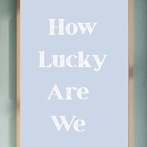 May include: A framed print with the words "How Lucky Are We" in white script on a light blue background. The frame is a light brown color, and the print is hanging on a light green wall.