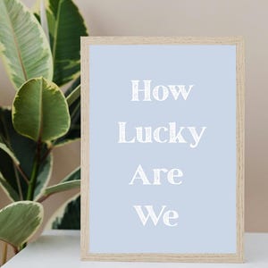 May include: A framed print with the words "How Lucky Are We" in white, centered on a light blue background. The frame is made of light-colored wood. A green plant is visible in the background.
