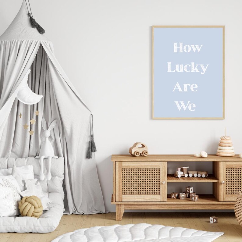May include: A light blue framed print with the text "How Lucky Are We" hangs on a white wall. A wooden cabinet with woven doors holds wooden toys. A gray canopy bed with a stuffed bunny and moon is in the corner.