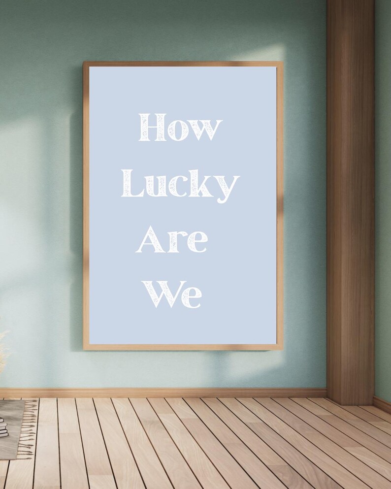 May include: A framed print with the words "How Lucky Are We" in white script on a light blue background. The frame is a light brown color, and the print is hanging on a light green wall.