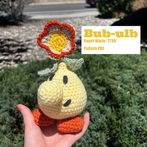 May include: A crocheted plushie of Bub-ulb from Paper Mario:TTYD. The plushie is yellow with a red flower on top and orange legs. The plushie is being held in a hand.