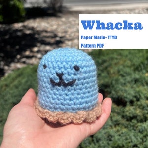 Peut inclure: Un Whacka bleu crocheté de Paper Mario- TTYD. La peluche a un visage souriant avec des détails brodés noirs et une base festonnée beige. Le texte "Whacka" et "Paper Mario- TTYD Pattern PDF" est dans un encadré blanc.