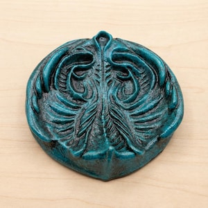 May include: A teal-colored, decorative dish with an ornate, symmetrical design. The dish has a raised, textured surface with a central, vertical element and swirling patterns. The dish is made of a solid material and sits on a light-colored wooden surface.