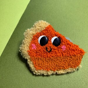 Peut inclure: Une part de tarte en crochet marron et orange avec un visage souriant, des yeux globuleux et des joues roses.