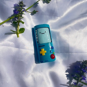 Puede incluir: Una figurita pintada a mano de una consola de videojuegos azul y amarilla con una cara sonriente. La consola tiene un botón amarillo con un signo más y botones rojo, verde y azul.