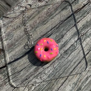 May include: A pink donut with colorful sprinkles hanging from a silver chain. The donut is on a clear acrylic surface.