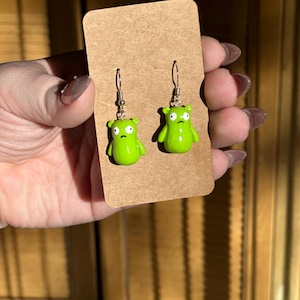 May include: A pair of green dangle earrings featuring a cartoon character with a surprised expression. The earrings are on a brown card.