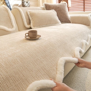 May include: Beige sofa with matching pillows and a coffee cup on the surface. The sofa has a textured fabric and is trimmed with a fluffy white material. The overall aesthetic is neutral and cozy.