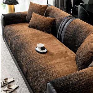 Peut inclure: Un canapé à motifs marron et noir avec des coussins assortis. Le canapé présente un aspect texturé et des détails en cuir noir. Un petit plateau avec une tasse et des boules décoratives est posé sur le canapé.