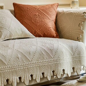 May include: Close-up of a sofa with decorative cushions and a patterned cover. The cushions are in shades of cream and rust-orange, with detailed embossed designs. The sofa cover has a repeating geometric pattern with a scalloped edge.