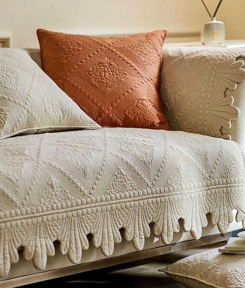 May include: Close-up of a sofa with decorative cushions and a patterned cover. The cushions are in shades of cream and rust-orange, with detailed embossed designs. The sofa cover has a repeating geometric pattern with a scalloped edge.