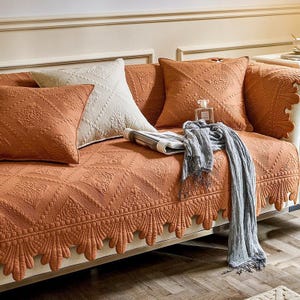 May include: A sofa with a rust-colored quilted cover and matching throw pillows. The cover features a detailed geometric pattern and a scalloped edge. A light gray scarf and a perfume bottle rest on the sofa.