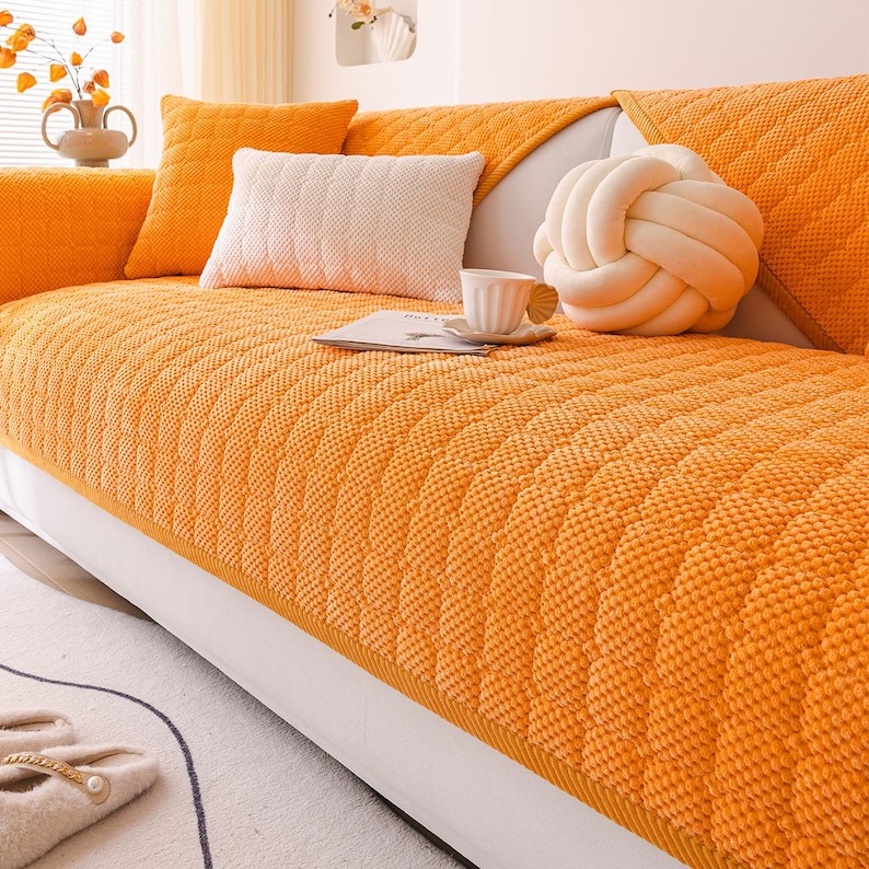K&ouml;nnte beinhalten: Eine orangefarbene Sofahusse mit einem strukturierten, gesteppten Muster. Das Sofa ist mit passenden orangefarbenen und wei&szlig;en Kissen sowie einem dekorativen, knotenf&ouml;rmigen Kissen ausgestattet. Eine wei&szlig;e Teetasse und Untertasse liegen auf einem kleinen Buch.