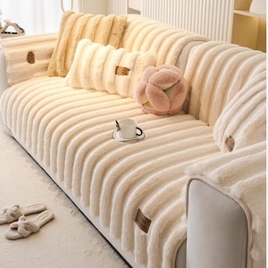 以下が含まれることがあります： クリーム色のソファ。 柔らかくふわふわした素材で覆われ、リブ編みのテクスチャが特徴です。 お揃いのクッションとピンクの装飾が施されています。 ふわふわのスリッパが床に置かれています。