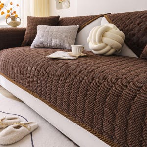 K&ouml;nnte beinhalten: Eine braune, gesteppte Sofahusse mit einem strukturierten Muster. Das Sofa ist mit dekorativen Kissen in Braun- und Graut&ouml;nen geschm&uuml;ckt. Ein wei&szlig;es, knotenf&ouml;rmiges Kissen und eine Tasse Kaffee sind ebenfalls zu sehen.