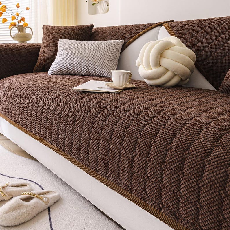 K&ouml;nnte beinhalten: Eine braune, gesteppte Sofahusse mit einem strukturierten Muster. Das Sofa ist mit dekorativen Kissen in Braun- und Graut&ouml;nen geschm&uuml;ckt. Ein wei&szlig;es, knotenf&ouml;rmiges Kissen und eine Tasse Kaffee sind ebenfalls zu sehen.