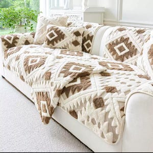 以下が含まれることがあります： 白のソファに、クリーム色のスローケットとクッションが置かれています。スローケットとクッションには、茶色の幾何学模様が繰り返されています。ソファは、窓のある明るい部屋に置かれています。