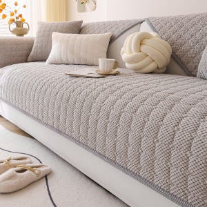K&ouml;nnte beinhalten: Eine hellgraue, gesteppte Sofahusse mit Rautenmuster, mit mehreren dekorativen Kissen in neutralen T&ouml;nen. Ein wei&szlig;es, knotenf&ouml;rmiges Kissen und eine kleine Teetasse auf einer Untertasse sind ebenfalls zu sehen. Das Sofa hat eine wei&szlig;e Basis.