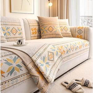 May include: A cream-coloured sofa adorned with patterned cushions and a matching throw. The cushions and throw display geometric patterns in yellow, blue, and orange. A pair of fluffy slippers with a black and white design are on the floor.