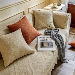 May include: A beige sofa with several decorative pillows in shades of beige and rust. The pillows feature a quilted diamond pattern with floral accents. A gray scarf and an open book rest on the sofa.