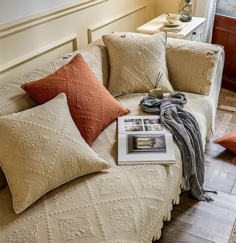 May include: A beige sofa with several decorative pillows in shades of beige and rust. The pillows feature a quilted diamond pattern with floral accents. A gray scarf and an open book rest on the sofa.