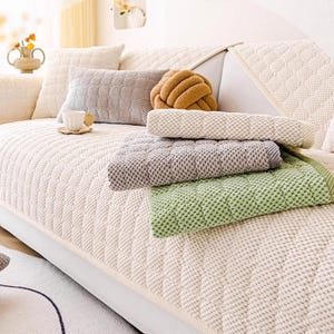K&ouml;nnte beinhalten: Ein cremefarbenes Sofa mit einem Steppmuster ist mit verschiedenen Kissen und Decken geschm&uuml;ckt. Ein graues rechteckiges Kissen, ein braunes knotenf&ouml;rmiges Kissen und strukturierte Decken in Gr&uuml;n, Grau und Creme sind zu sehen.