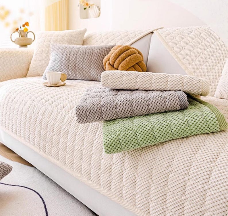 K&ouml;nnte beinhalten: Ein cremefarbenes Sofa mit einem Steppmuster ist mit verschiedenen Kissen und Decken geschm&uuml;ckt. Ein graues rechteckiges Kissen, ein braunes knotenf&ouml;rmiges Kissen und strukturierte Decken in Gr&uuml;n, Grau und Creme sind zu sehen.