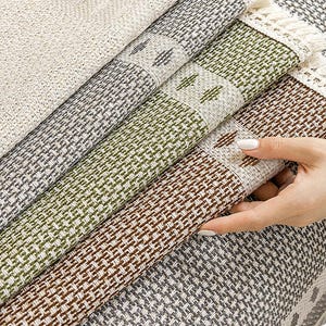 Op de afbeelding: Close-up van verschillende opgerolde geweven tapijten in tinten beige, grijs, groen en bruin. Elk tapijt heeft een textuurpatroon en een decoratieve rand. De tapijten zijn gemaakt van een duurzaam materiaal, geschikt voor woondecoratie.