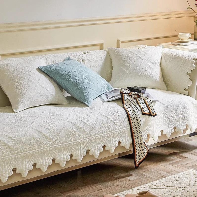 May include: A white quilted sofa cover with a scalloped edge, adorned with matching throw pillows. A light blue pillow and a patterned scarf add color. The sofa is in a well-lit room with a neutral color palette.