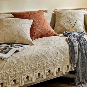 May include: A beige sofa with several decorative pillows and a patterned sofa cover. The pillows are in shades of beige and rust, with intricate quilted designs. A gray scarf with fringe and a magazine are also on the sofa.