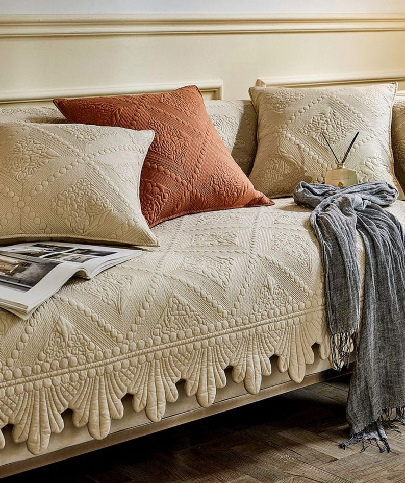 May include: A beige sofa with several decorative pillows and a patterned sofa cover. The pillows are in shades of beige and rust, with intricate quilted designs. A gray scarf with fringe and a magazine are also on the sofa.