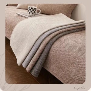 May include: A stack of folded blankets in shades of beige, gray, and brown rests on a textured sofa. A checkered mug and a book are on the sofa. The word "Comfortable" is written in cursive in the bottom right corner.