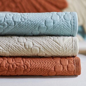 May include: A stack of three folded, quilted blankets in shades of blue, cream, and rust. Each blanket features a repeating pattern of circles and diagonal stitching. The blankets are neatly stacked, showcasing their texture and color variations.
