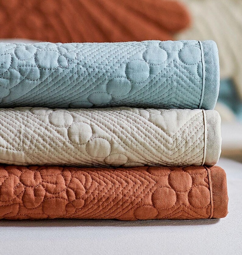 May include: A stack of three folded, quilted blankets in shades of blue, cream, and rust. Each blanket features a repeating pattern of circles and diagonal stitching. The blankets are neatly stacked, showcasing their texture and color variations.